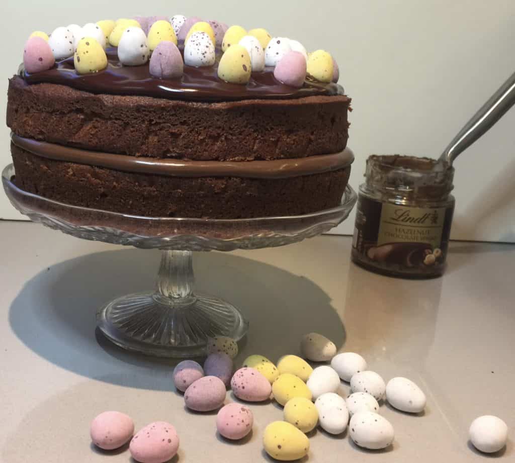 Chocolate Cake with Lindt Hazelnut chocolate spread filling. Decorated with mini sugar coated eggs. Displayed on a glass cake stand. 