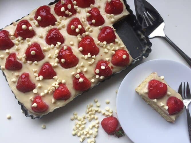 overhead shot of vanilla sheet cake topped with a white chocolate frosting and fresh strawberries , fresh strawberries and white chocolate chips.