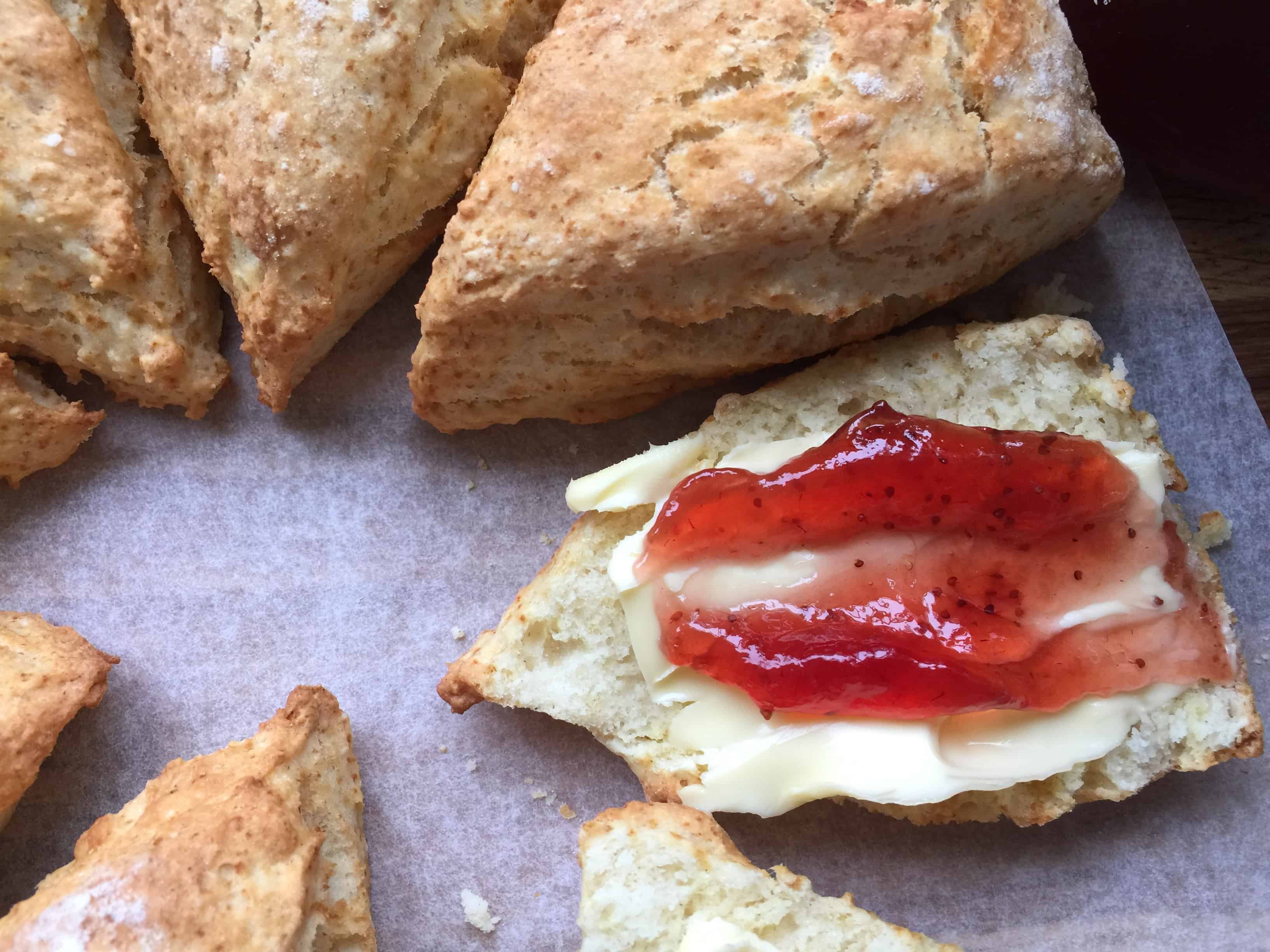 A round of baked scones. One sliced with butter and jam.